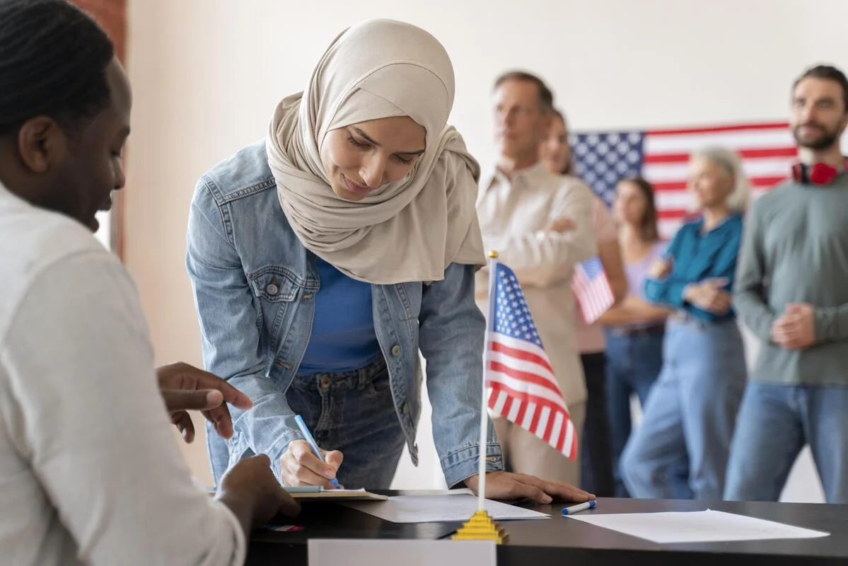A woman in a hijab signing paperwork at an immigration office, with people waiting in line and an American flag in the background, representing the asylum process in California.
