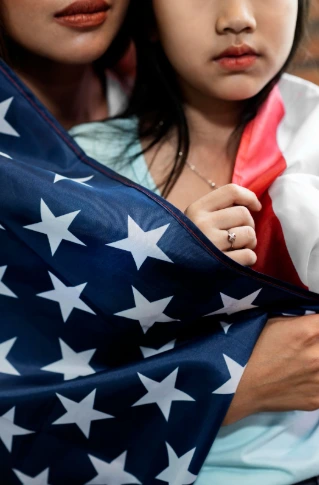A woman holds a young girl, enveloped in an American flag, representing the ideals of unity and the significance of citizenship. Can Trump End Birthright Citizenship?