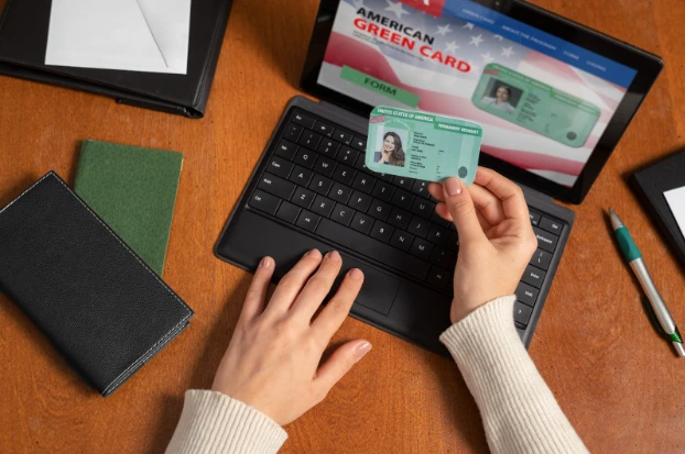 A woman's hands typing on a laptop, accompanied by a green card and passport, symbolizing immigration processes.How Long Does It Take to Renew a Green Card?